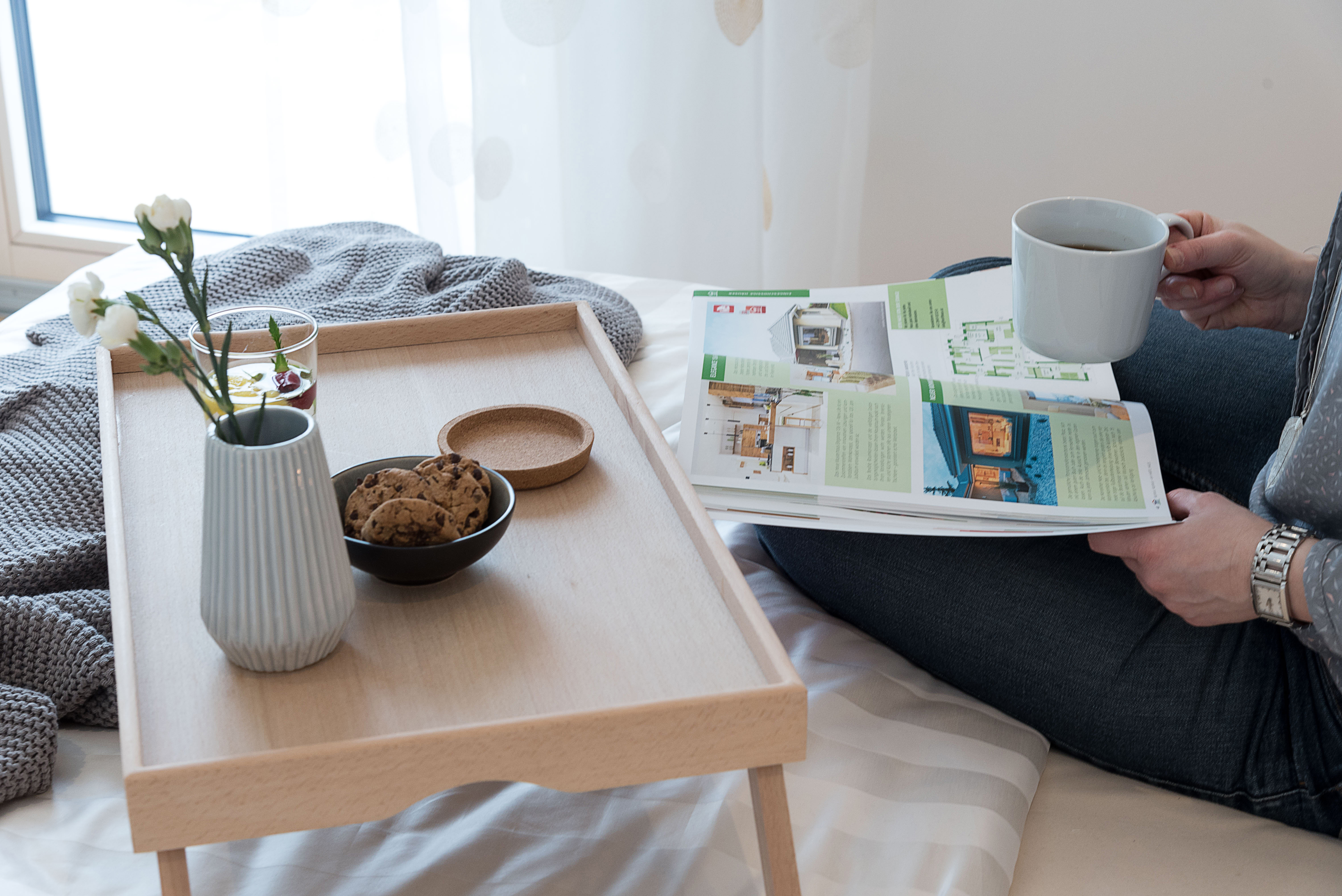 Frau liest Hartl Haus Katalog im Bett. Tablet aus Holz mit Keksen und Kaffee steht auf dem Bett