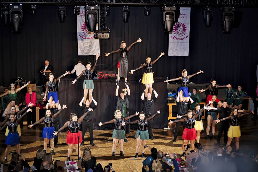 Gruppenfoto einer Tanzgruppe auf einer Bühne mit bunten Röcken und schwarzem Oberteil bei einer Aufführung.