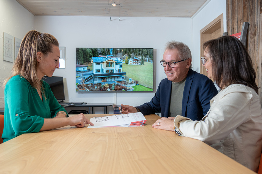 Drei Personen sitzen an einem Tisch und besprechen Baupläne mit einem Bildschirm im Hintergrund, der ein Haus und eine grüne Landschaft zeigt.
