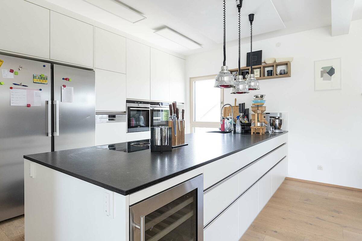 Küche mit Kücheninsel und großem Edelstahl-Kühlschrank. Weiße Möbel gepaart mit schwarzer Arbeitsplatte