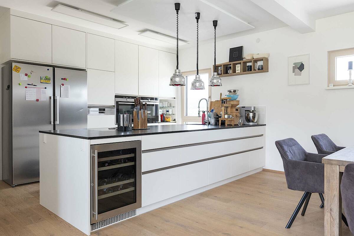Küche mit Kücheninsel und großem Edelstahl-Kühlschrank. Weiße Möbel gepaart mit schwarzer Arbeitsplatte. Wärmelampen über der Kochinsel