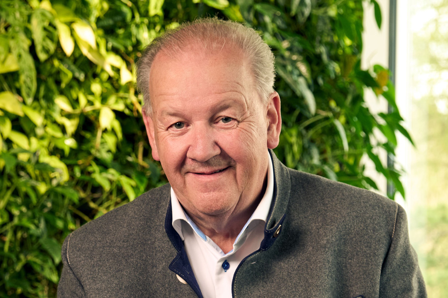 Älterer Mann mit grauem Jackett und weißem Hemd vor grünem Pflanzenhintergrund.