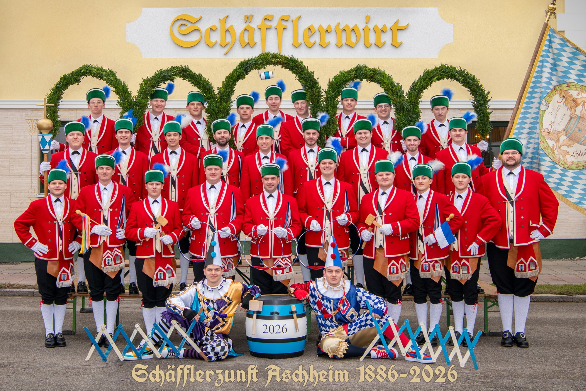 Gruppenfoto von Männern in traditionellen roten und grünen Trachten vor dem Schild 'Schäfflerwirt' mit dem Text 'Schäfflerzunft Aschheim 1886-2026'.