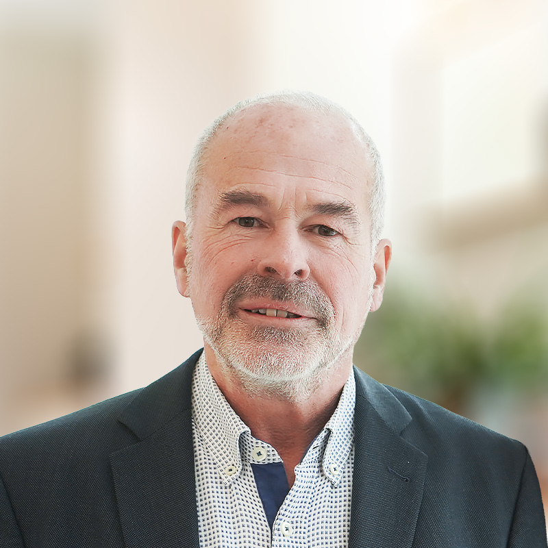 Älterer Mann mit kurzem grauem Haar und Bart, in kariertem Hemd und dunklem Sakko, vor hellem unscharfem Hintergrund.