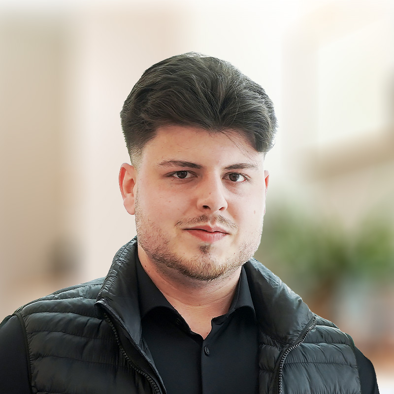 Porträt eines jungen Mannes mit dunklen Haaren in einem schwarzen Hemd und einer schwarzen Steppweste vor unscharfem Hintergrund.