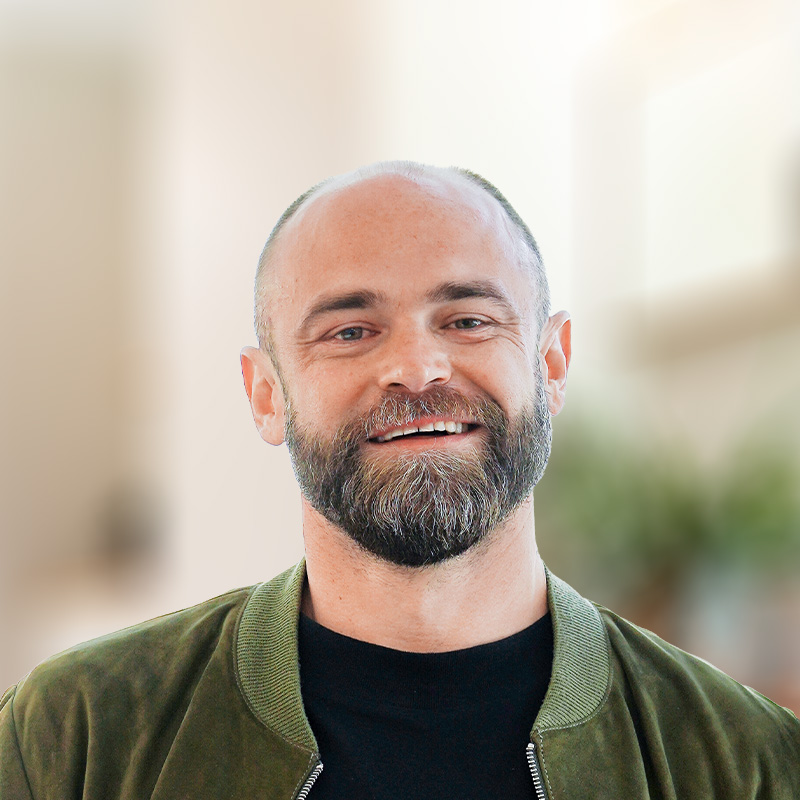 Lächelnder Mann mit Glatze und dunklem Bart trägt eine olivgrüne Jacke und ein schwarzes T-Shirt vor einem hellen, verschwommenen Hintergrund.